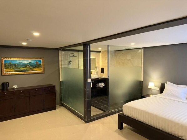 A modern hotel room with a bed, a dark wooden dresser, a glass partition, a bathroom with a mirror, and a painting on the wall. - Kooters Guesthouse Bali (Legian)