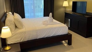 A modern hotel room with a large bed, a flat-screen TV, a bedside table with a lamp, and a view of the city through the window. - Kooters Guesthouse Bali (Legian)