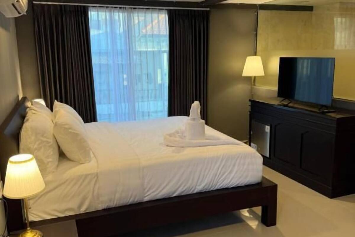 A modern hotel room with a large bed, a flat-screen TV, a bedside table with a lamp, and a view of the city through the window.