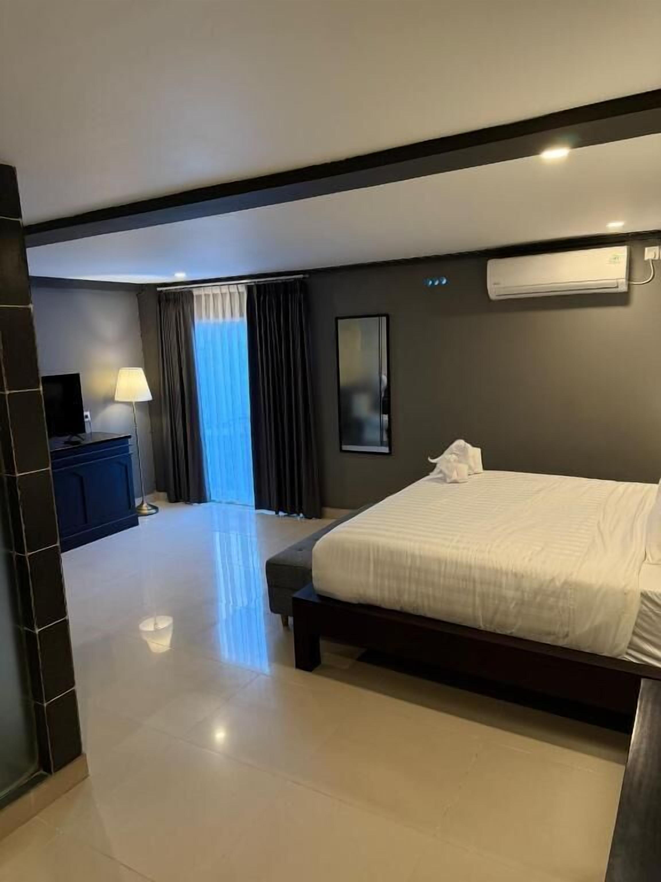 A hotel room with a large bed, a desk with a lamp, a TV, and an air conditioning unit.