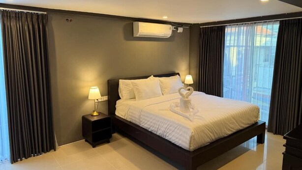 A modern hotel room with a large bed, two bedside tables with lamps, a wall-mounted air conditioner, and a view of the city through large windows. - Kooters Guesthouse Bali (Legian)