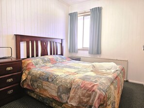 A bedroom with a wooden headboard, a quilted bedspread, a bedside table with drawers, a window with curtains, and a radiator. - Room 01 : Cozy Studio 4 Guests Central Walk / In The Heart Of Jindabyne (Jindabyne)
