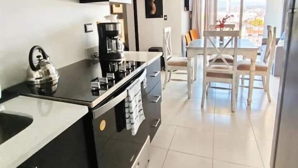 Una cocina moderna con armarios negros, un hervidor de acero inoxidable y una cafetera. Hay una zona de comedor con una mesa y sillas.