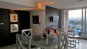 Un comedor moderno con una mesa grande y sillas, un televisor de pantalla plana montado en la pared, y vistas de la ciudad a través de amplios ventanales.