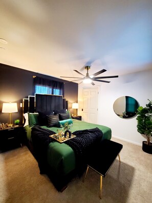 A bedroom with a bed, bedside tables, a ceiling fan, a round mirror, and a potted plant. - GOLDEN RAVEN House (Indio)