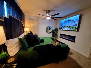 A hotel room with a bed, a ceiling fan, a TV displaying a Ferris wheel, and a small plant. - GOLDEN RAVEN House (Indio)
