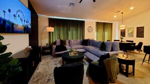 A modern living room with a sectional sofa, a dining table with chairs, a ceiling fan, and a large TV screen displaying a scenic image. - GOLDEN RAVEN House (Indio)
