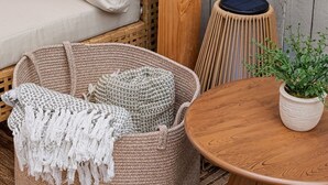 Un panier en osier avec une couverture à franges blanches, une table ronde en bois sur laquelle est posée une plante en pot, et un tabouret en osier avec un plateau noir.