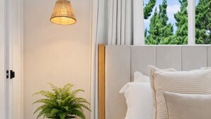 Une chambre à coucher avec un lit, une table de chevet ornée d’une plante, une lampe, et une vue sur les arbres par la fenêtre.