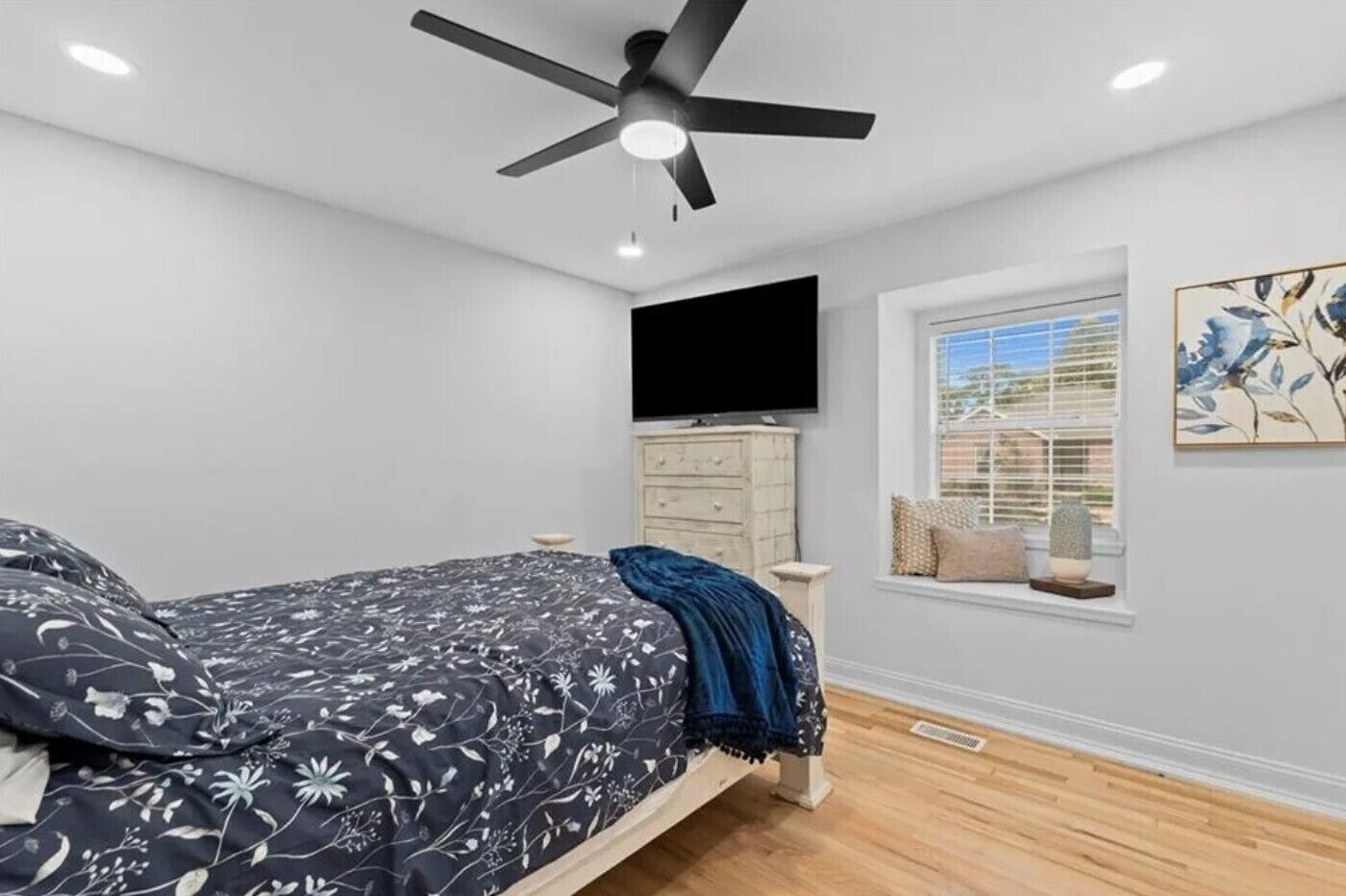 Een slaapkamer met een bed, een plafondventilator, een televisie, een raam met jaloezieën en een schilderij aan de muur.
