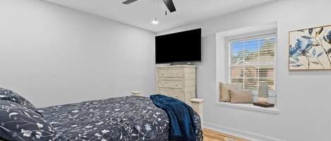 Een slaapkamer met een bed, een plafondventilator, een televisie, een raam met jaloezieën en een schilderij aan de muur.