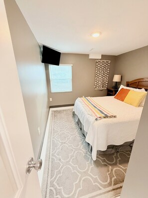 A bedroom with a bed, a TV mounted on the wall, a window with blinds, a patterned rug, and a nightstand with a lamp. - Cozy Guest Suite near Downtown Lee's Summit (Lee's Summit)