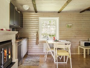 En mysig stuga med träpanel på väggarna, en öppen spis, ett matbord med stolar och ett fönster med utsikt över träd.