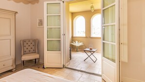 A room with a bed, a chair, a door leading to a balcony with arched windows, and a small table with a vase.