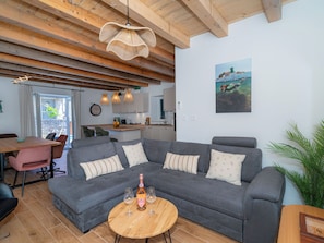 Un salon chaleureux avec un canapé d’angle gris, une table basse en bois et un coin repas agrémenté d’une chaise rose.