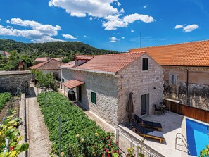 Un bâtiment en pierre avec un toit en tuiles, une piscine et un jardin où poussent différentes espèces de plantes.