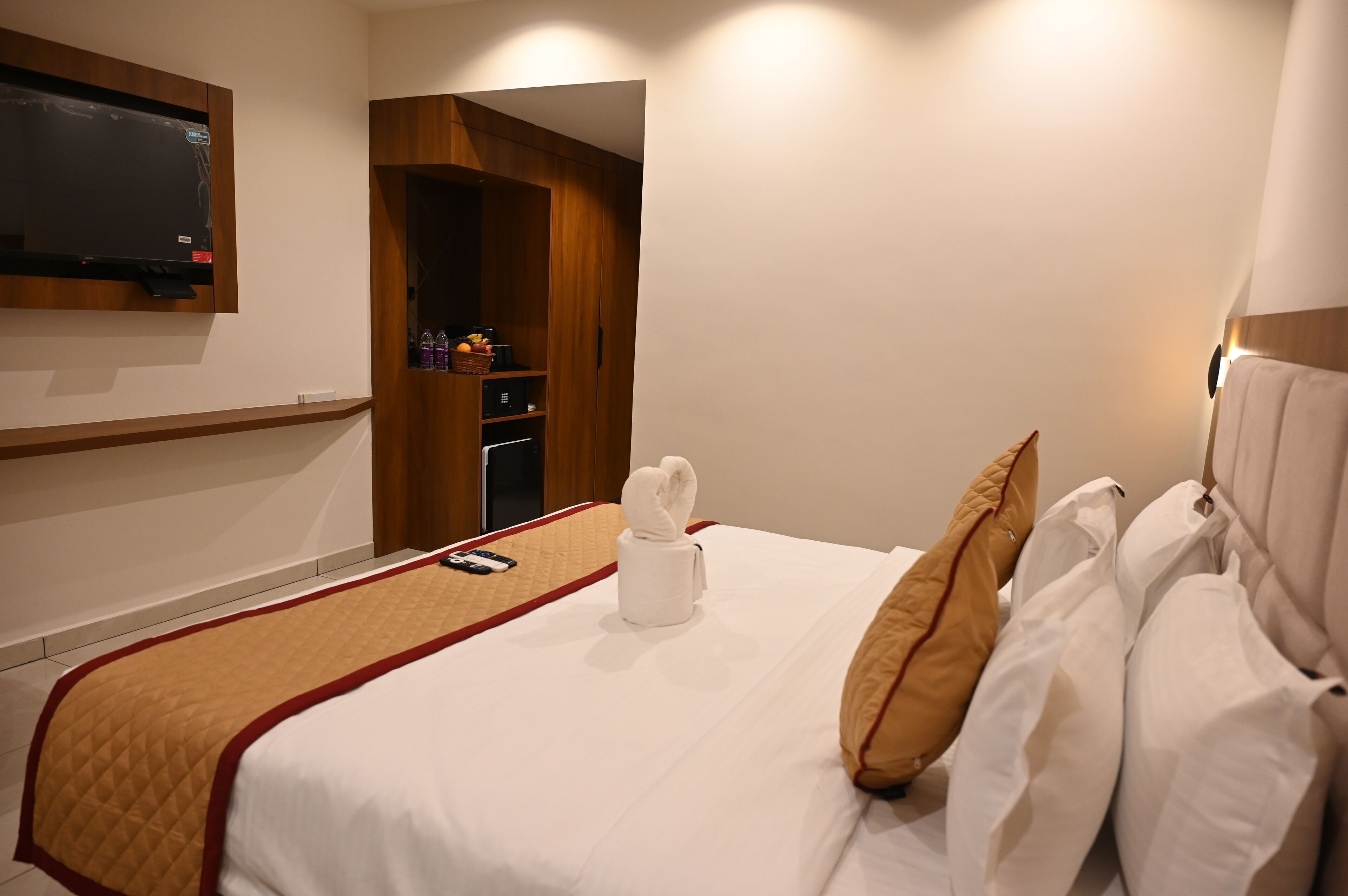 A hotel room with a bed, a TV mounted on the wall, a wooden wardrobe, and a towel on the bed.