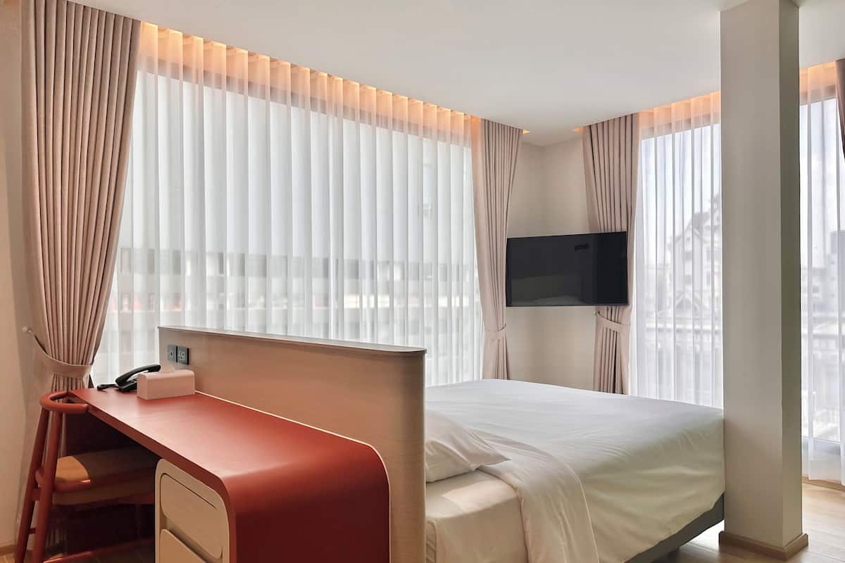 A hotel room with a bed, a desk, a chair, a TV, and large windows with curtains.