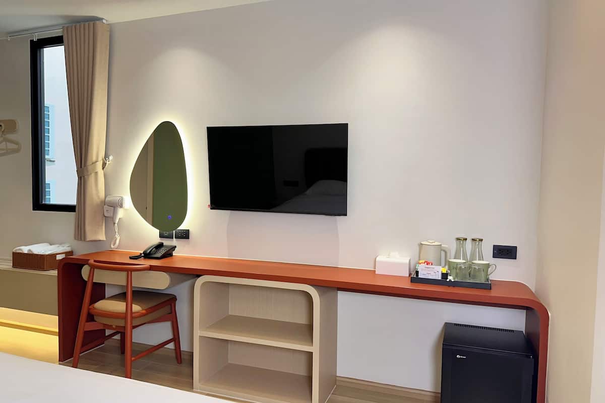A hotel room with a wooden desk, a chair, a flat-screen TV mounted on the wall, a small shelf, a bedside table, and a black refrigerator.