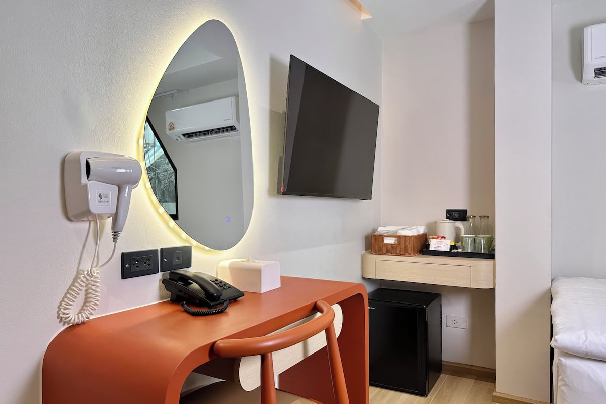 A modern hotel room with a desk, chair, hair dryer, phone, mirror, TV, and a small trash bin.
