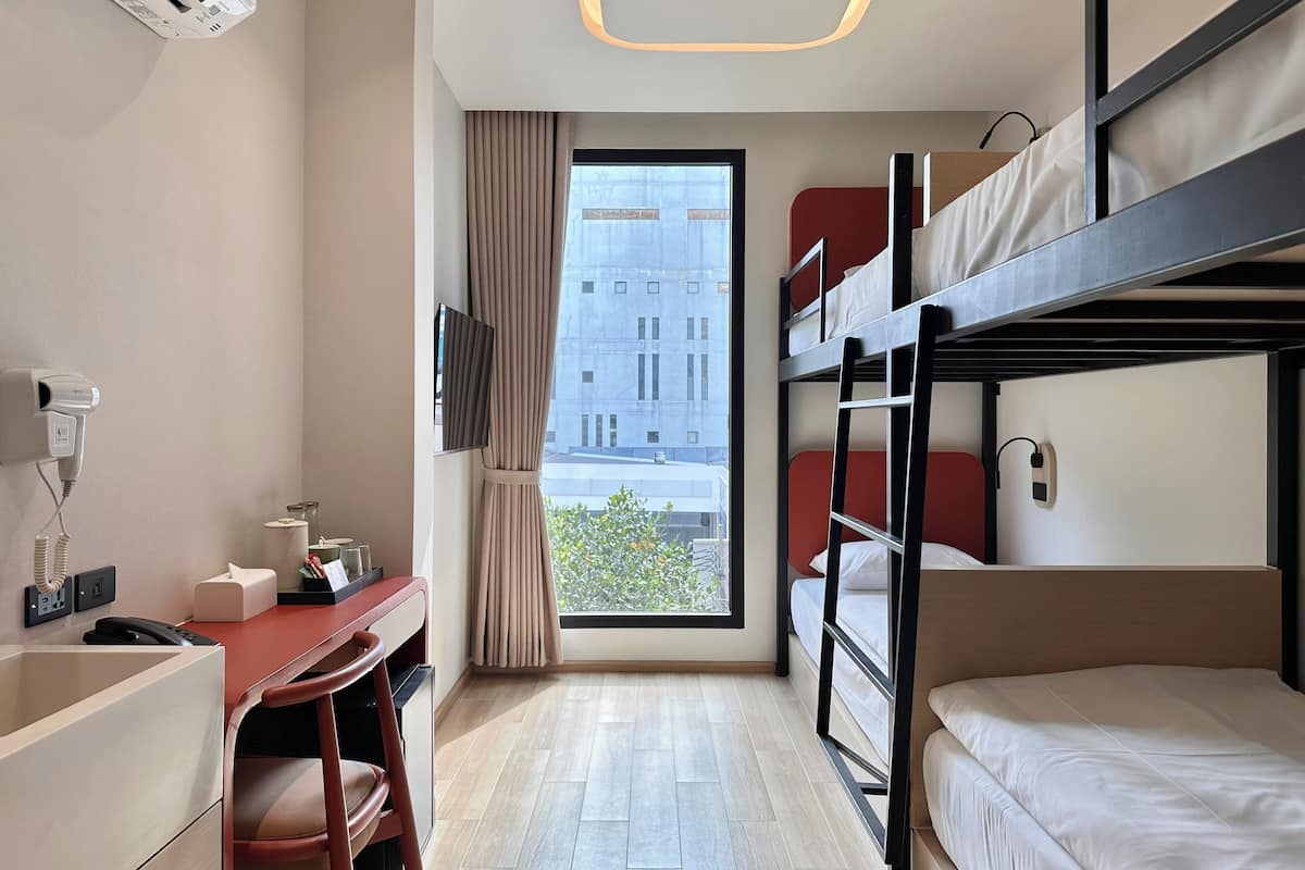 A compact hotel room with bunk beds, a desk with a chair, a hairdryer, and a window with curtains.