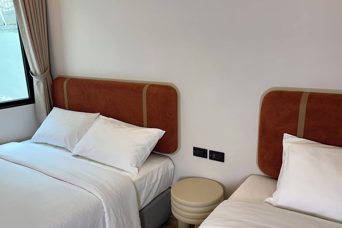 A hotel room with two beds, each with white bedding and brown headboards. There is a small stool between the beds, and a window with curtains on the left side.