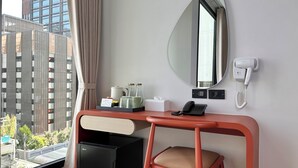 Hotellihuone, jossa on työpöytä, tuoli, peili, puhelin, minijääkaappi ja ikkuna, josta on näkymä rakennuksiin.