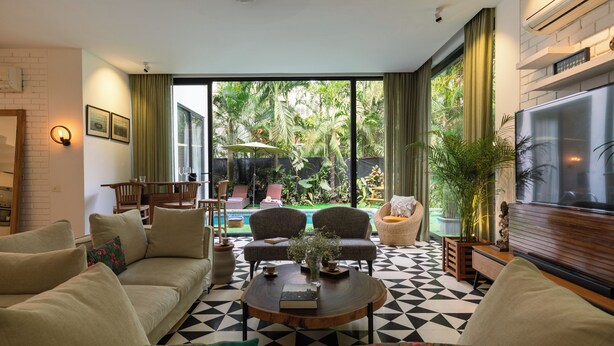 Ein Wohnzimmer mit einer Couch, Sesseln, einem Couchtisch und einem Fernseher. Ein großes Fenster bietet Blick auf Grünfläche.