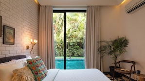 Ein Schlafzimmer mit einem großen Bett, Blick auf einen Swimmingpool, einer Topfpflanze und einem kleinen Tisch mit Buch und Tasse.