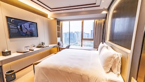 Quarto de hotel com uma cama grande, uma mesa com computador, uma TV fixada na parede e vista para a cidade pela janela.