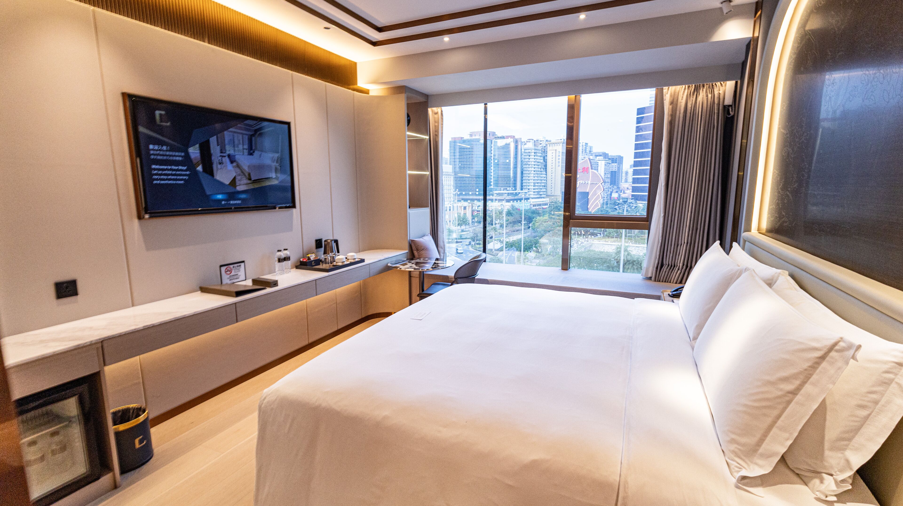A modern hotel room with a large bed, a TV mounted on the wall, a window with a city view, and a small table with amenities.