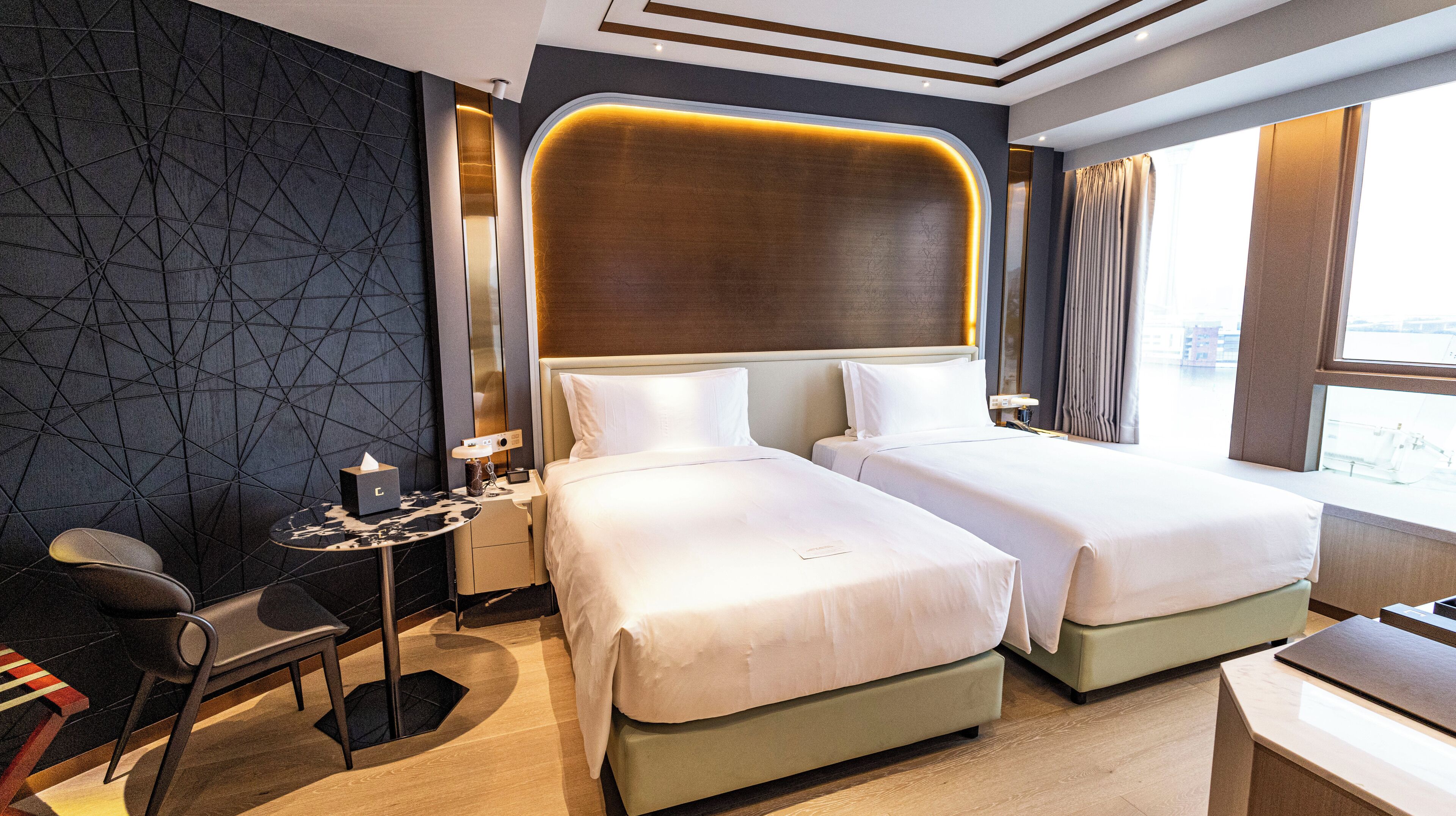 A hotel room with two beds, a small table with a lamp, a chair, and a view of the city through the window.