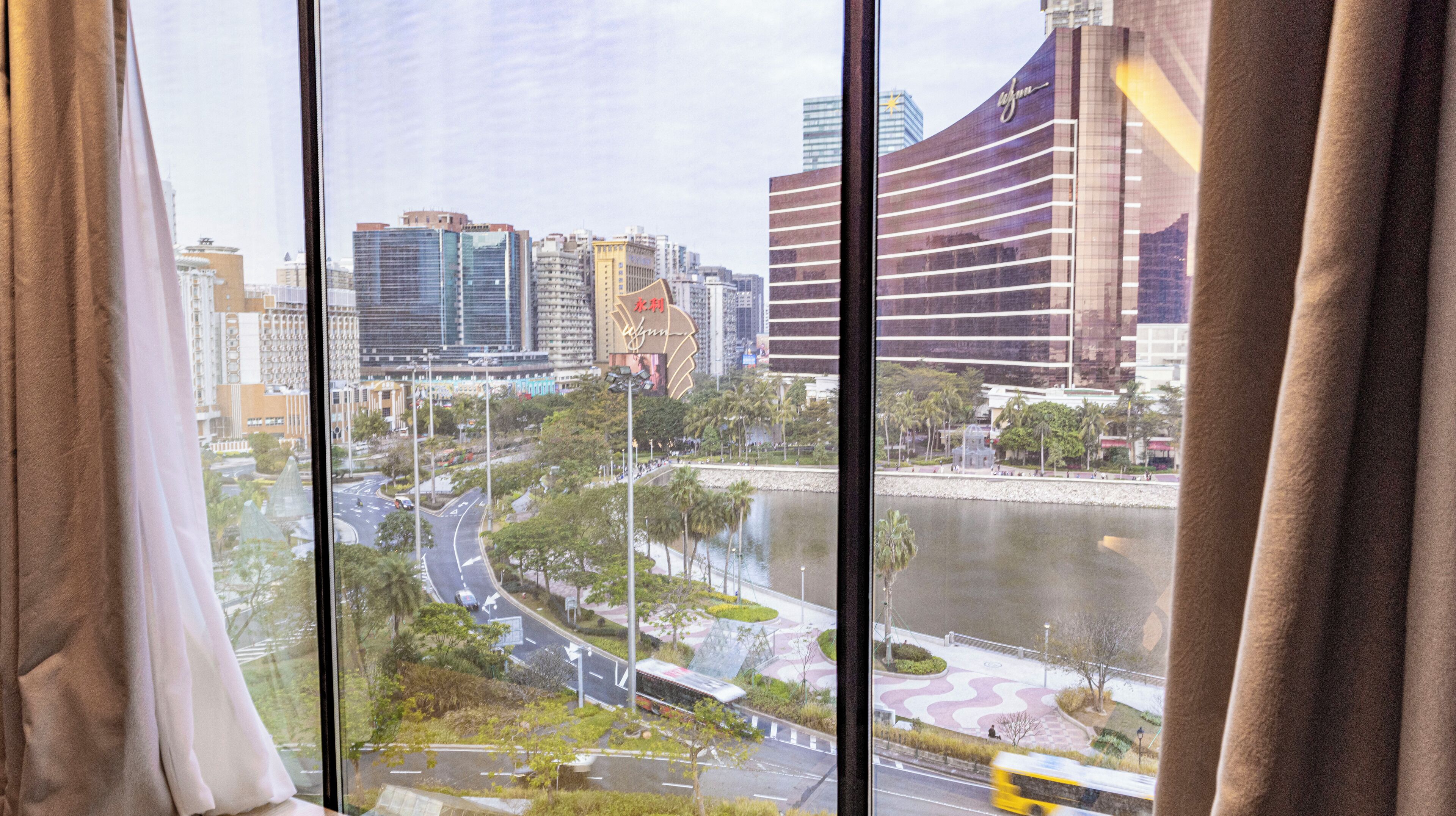 A view of a cityscape through a window with curtains, featuring modern buildings, a river, and a landscaped area with trees and a pathway.
