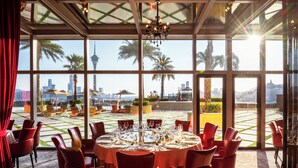 Restaurante com mesas redondas preparadas para refeições, amplas janelas oferecendo vista para palmeiras e horizonte urbano.