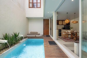 A modern pool area with a wooden deck, a seating area with a cushion, and a kitchen visible through glass doors. - Sandwave Villa by Betterplace (Ungasan)