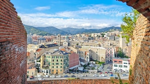 Paesaggio urbano visibile attraverso un arco in mattoni, con un mix di edifici storici e moderni, una piazza centrale e montagne sullo sfondo.