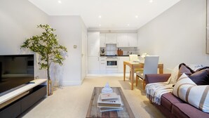 A modern living room with a flat-screen TV, a coffee table with books and a decorative item, a sofa with cushions, a dining area with a table and chairs, and a kitchen in the background. - Hertford Flat 15 (London)