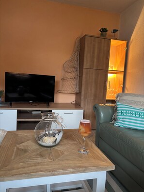 Un salon avec un téléviseur, un saladier en verre contenant divers objets, un verre à vin, un canapé vert et un meuble en bois.