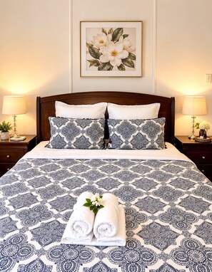 Un lit bien fait, recouvert d’une couvre-lit à motifs, avec deux oreillers et, au-dessus, un tableau floral encadré.