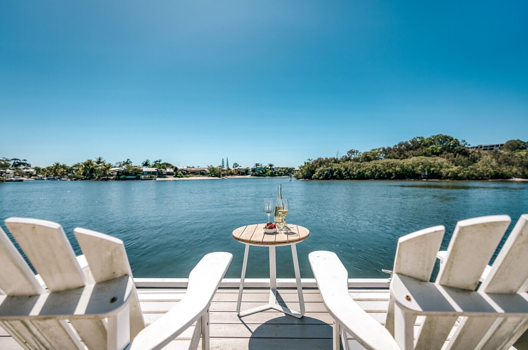 Une terrasse avec deux chaises blanches disposées face à une table sur laquelle se trouvent une bouteille et des verres, donnant sur une étendue d’eau calme sous un ciel dégagé.