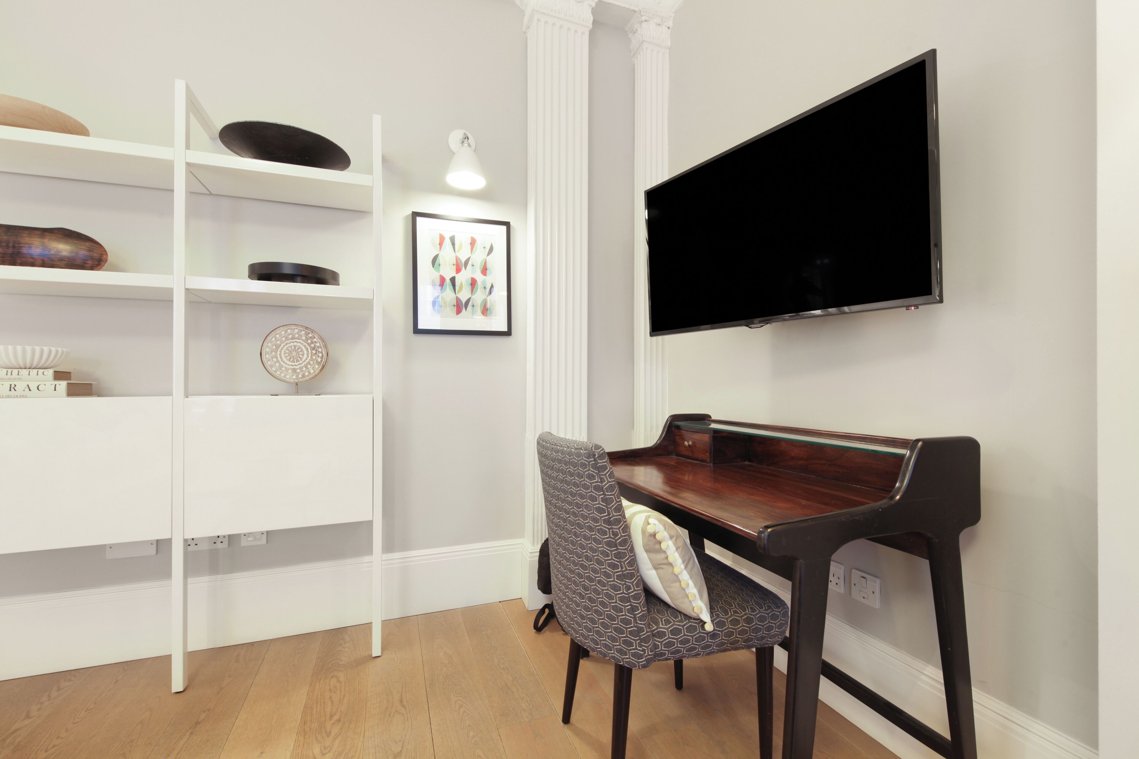 Una habitación con un piano, un televisor montado en la pared, una estantería blanca con objetos decorativos y una silla estampada.
