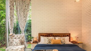 Une chambre avec un mur en briques, un grand lit avec de nombreux oreillers, une chaise sur laquelle est posé un chapeau, et une vue sur la verdure à l’extérieur.