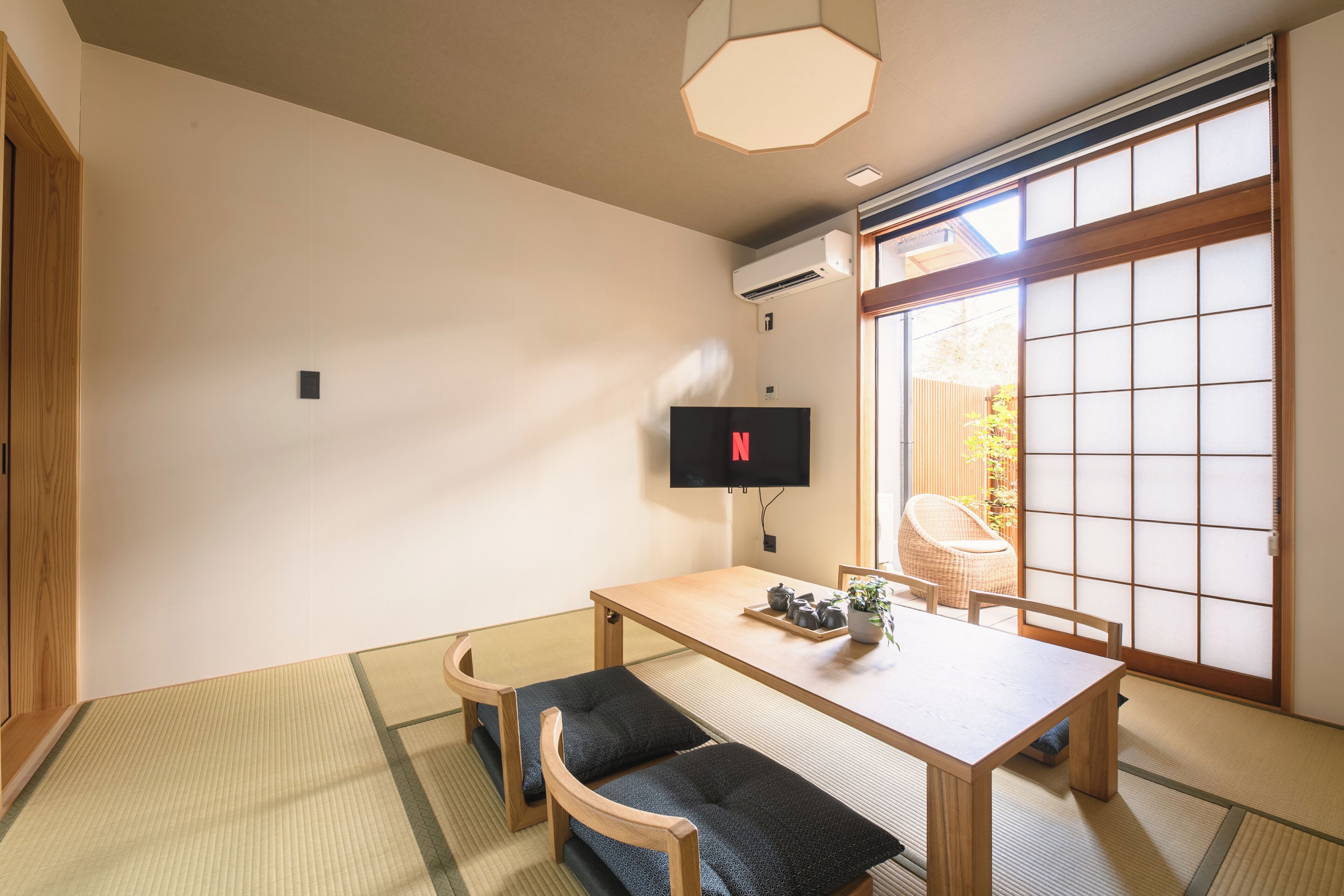 Quarto japonês tradicional com piso de tatame, uma mesa de madeira baixa com almofadas, uma televisão e uma porta deslizante que dá para um jardim.
