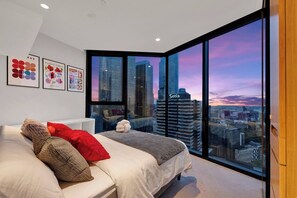 Une chambre moderne avec un grand lit, une vue sur les gratte-ciel de la ville et des œuvres d’art encadrées accrochées au mur.