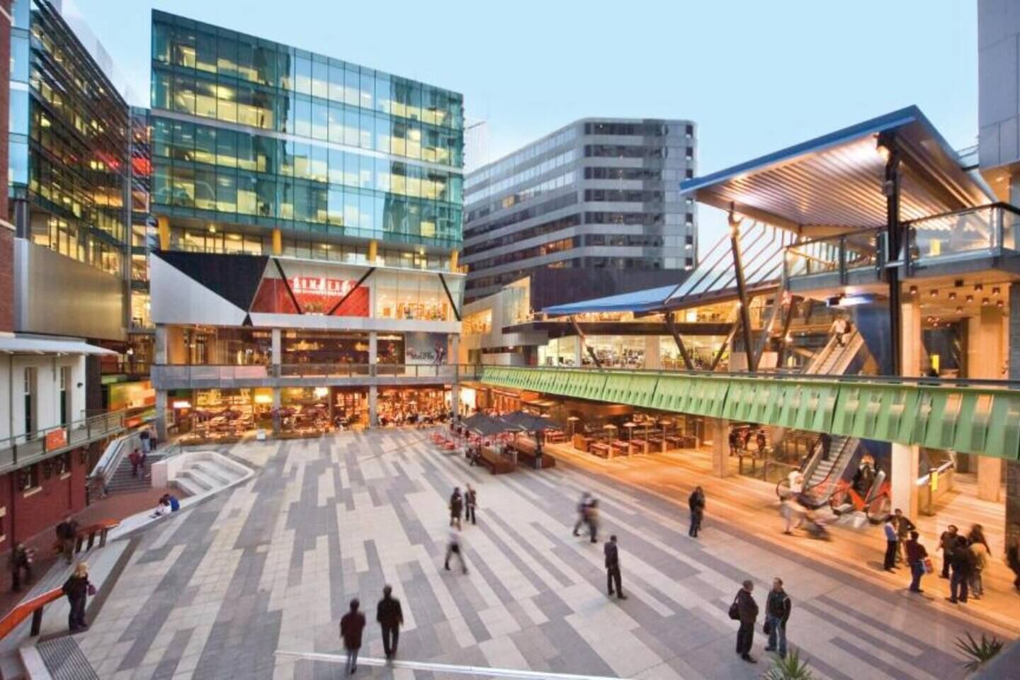 A modern urban plaza with a glass building, a covered walkway, and a central open space with seating and people.