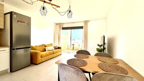 A modern kitchen with a refrigerator, a yellow sofa, a dining table with woven placemats, and a large window with curtains. - Welooveyou Baltico your place to discover the island (Costa Calma (incl. Urbanización Cañada del Río))