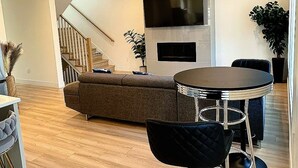 Una sala de estar moderna con un sofá, una mesa redonda con sillas, un televisor de pantalla plana montado en la pared y una escalera que sube a otro nivel.