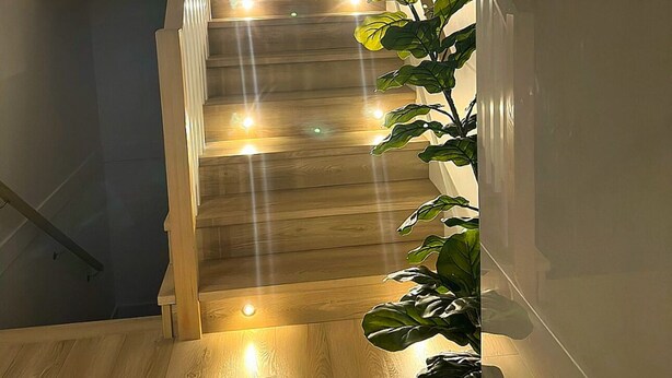 Una escalera con escalones de madera e iluminación empotrada, que conduce a una zona de descanso con un televisor de pantalla plana montado en la pared.