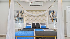 Uma cama de dossel com estrutura dourada, roupa de cama azul e preta e um dossel branco. Há duas mesas de cabeceira com luminárias e arte abstrata nas paredes, de ambos os lados da cama.