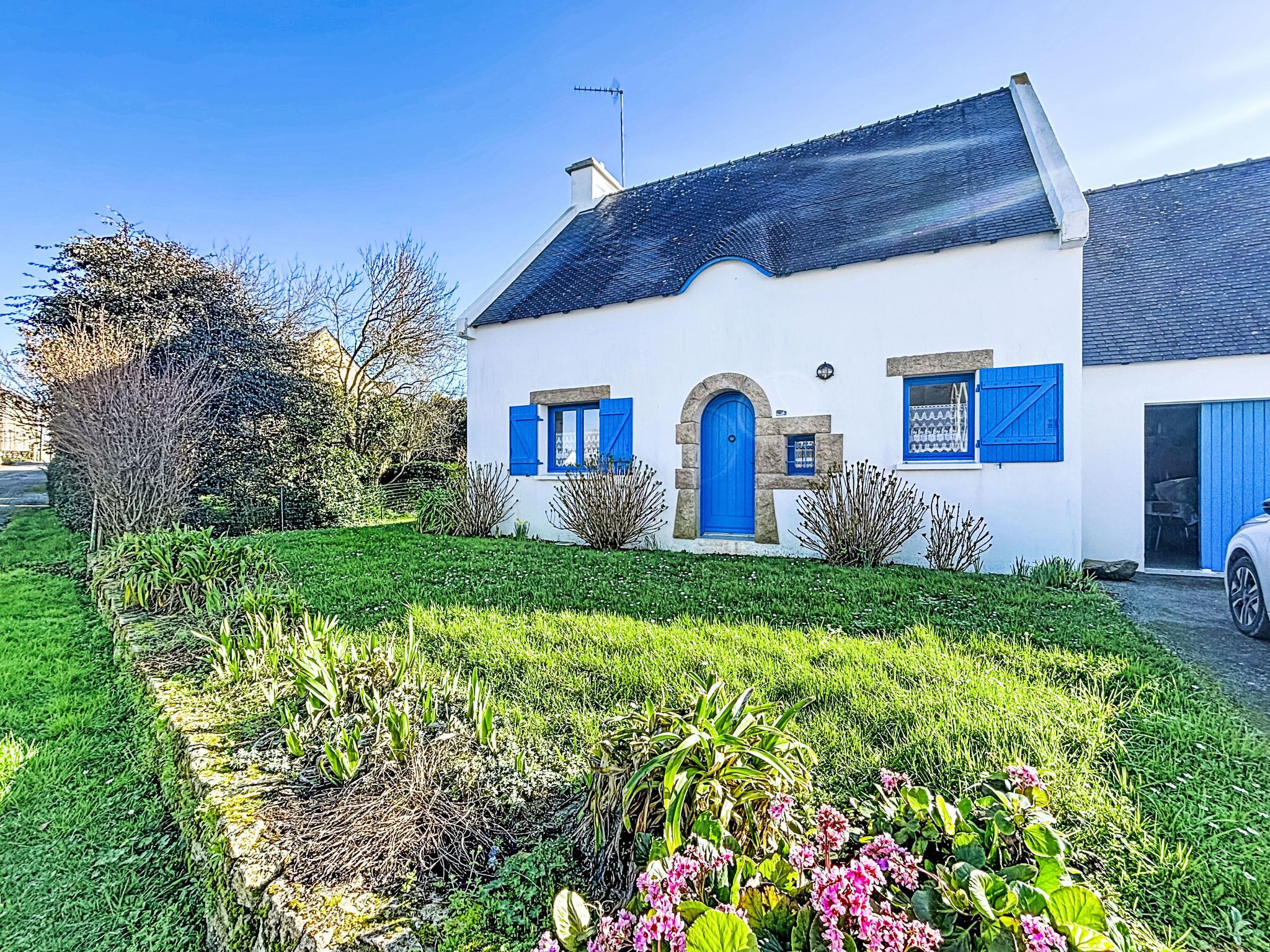 Una casa blanca con contraventanas y puerta azules, rodeada de jardines cuidados y un cielo despejado.
